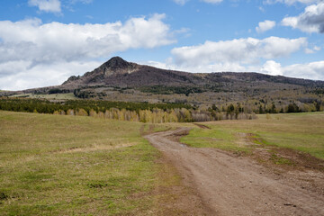 Fototapeta premium Mountain landscape, peak rises above the forest, mountain against the blue sky, clouds float, country road stretches across the field, journey trekking in nature