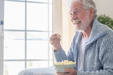 Senior 70s man seated on sofa eating popcorn, leisure and people concept - happy bearded senior man...