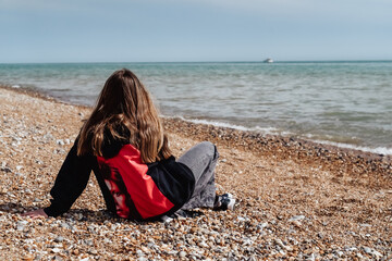 A lonely girl in a black sweater with long hair sits on the beach. A girl sits on the seashore and looks at the sea.
