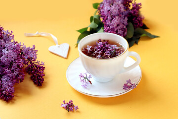Start your day with a cup of fragrant lilac tea: a white cup of tea, lilac branches, a ceramic heart on a yellow background, space for text