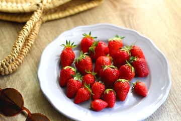 Lilac plate full of fresh strawberries, straw bag and retro sunglasses on wooden table. Selective focus.