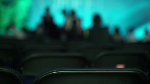 Public event. Concert, lecture, talk. Close-up of the backs of chairs arranged in even rows. Silhouettes of chairs on a background of colored light. Blurry silhouettes of people in the distance.