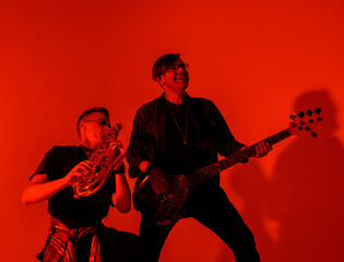 Obraz premium Young caucasian saxophonist playing saxophone against guitarist playing bass guitar against red light