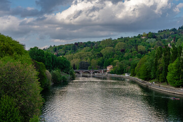 Fototapeta premium Natural landscape of the river Po in Torino on a spring afternoon. Green and leafy trees. Cloudy sky. And an old bridge that crosses the river.