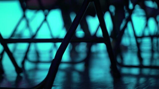An abstract geometric pattern made of even rows of chairs. Silhouettes of chairs on a background of colored light. Blurry silhouettes of people in the distance. Public event. Concert, lecture, talk.