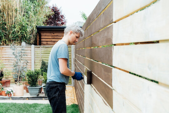 Young man worker paints with a roller a wooden board fence in the garden. DIY, Do it yourself concept. House improvement. Home renovation and refurbishment. Selective focus, copy space