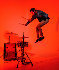 Young drummer jumps on his drums and holds drumsticks in his hands on a red light background