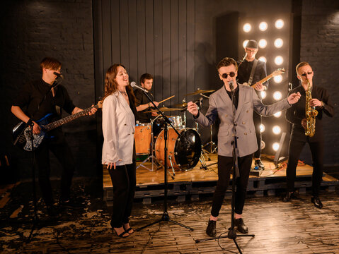 Two Caucasian vocalists singing against the backdrop of a band in the garage with yellow lights