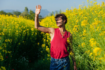 Naklejka na ściany i meble Young gay man with curly hair and raised hand, protecting from the sunlight, standing oilseed rapes. Yellow flower field at spring.