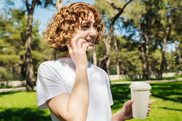 Beautiful redhead woman wearing white tee talking on phone and holding takeaway coffee cup looking away while standing on green city park, outdoors.