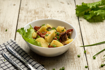 Stewed potatoes in a white cup on a white wooden table, cutlery and a checkered towel, food photo for a magazine and space for text, potatoes with meat and tomatoes, onions and lettuce