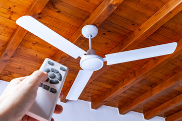 A man uses a remote control to turn on a white ceiling fan mounted in a house with wooden ceilings. - obrazy, fototapety, plakaty