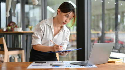 Female financial assistant looking the details on laptop screen and writing on clipboard