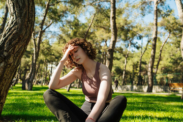 Attractive redhead woman sitting on the grass at the park with strong headache. Touching her temples feeling stress or pain. Stress or headache concept. Copy space.