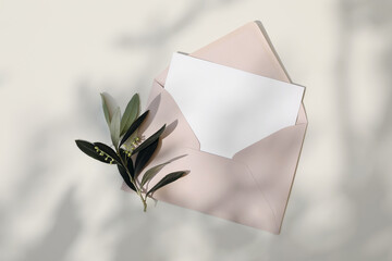 Summer Mediaterranean wedding stationery still life. Blank greeting card, imvitation mockup. Blush pink envelope in sunlight, beige table background in sunlight. Green olive tree branches. Flat lay