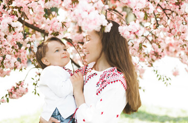 Fototapeta premium Little boy on hands of mother. Woman playing with child outside in blooming spring garden. Portrait of family of two people. Spring landscape background.