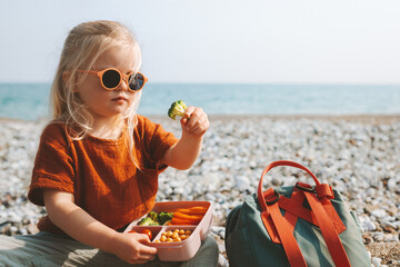 Child girl eating broccoli with lunch box picnic on beach vegan healthy food travel lifestyle outdoor summer vacations kid with backpack and lunchbox