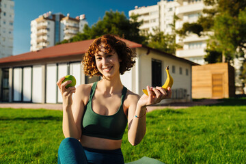 Beautiful redhead woman wearing green sports bra and blue yoga pants standing on city park, outdoors holding green apple and banana while sitting on yoga mat. Happy woman looking at the camera.