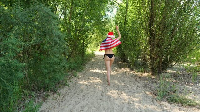Rear view of a girl in a black swimsuit walking along the sand between the trees. Brunette woman with USA flag. Independence Day. holiday relaxation