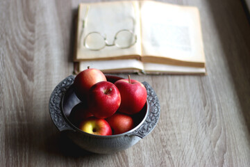 Silver bowl filled with red apples, open book and reading glasses on the table. Selective focus.