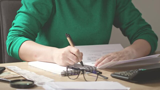 unrecognizable female hands writing budget numbers notes after home utility bills counting. checks, money, calculator, glasses, papers lying on desk besides woman in green sweatshirt. concept finance 
