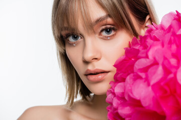 young woman with bangs and rhinestones under blue eyes near pink flower isolated on white
