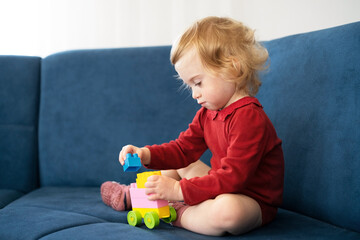 Beautiful cute caucasian baby girl, toddler with curly blond hair playing with erector set, sitting on sofa, serious infant, kid, having fun, spending time indoors at home