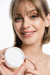 joyful young woman with bangs and cream on cheek holding container isolated on white