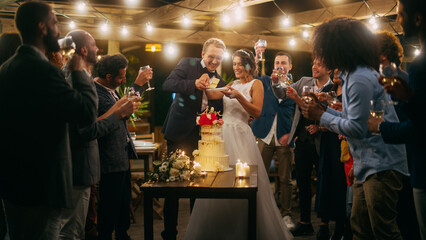 Beautiful Bride and Groom Celebrate Wedding at an Evening Reception Party with Multiethnic Friends. Married Couple Standing at a Dinner Table, Kiss and Cut Wedding Cake.