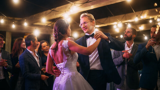 Beautiful Bride in White Dress and Groom in Stylish Black Suit Celebrate Wedding at an Evening Reception Party. Newlyweds Dancing at a Venue with Best Multiethnic Diverse Friends.