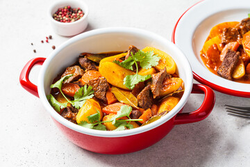 Beef stew with potatoes, carrots in tomato sauce in red pan, gray background.