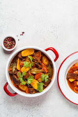 Beef stew with potatoes, carrots in tomato sauce in red pan, gray background.