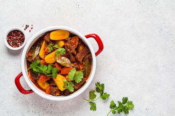 Beef stew with potatoes, carrots in tomato sauce in red pan, gray background.