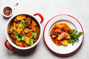 Beef stew with potatoes, carrots in tomato sauce in red pan, gray background.