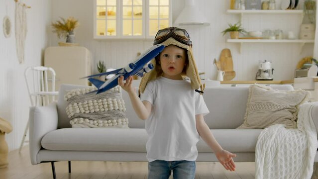Happy child in aviapilot hat is holding toy airplane in his hands, circling with an airplane and dreaming of flying in clouds. Child aspires to fly high in clouds playing with an airplane at home.