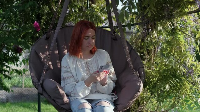 Young girl uses smartphone and swinging, sitting in hanging chair in backyard on a sunny day. Ukrainian female in national traditional clothes vyshyvanka 