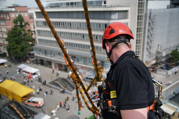Ein Höhenretter der Feuerwehr seilt sich von einem Hochhaus in der Innenstadt während eines Einsatz ab, unten warten Rettungswagen und weitere Helfer für den Notfall