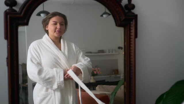 Young overweight woman is putting on white bathrobe and standing in home interior spbd. Closeup view of beautiful oversized female shows underwear on her body and puts on soft robe, looks in mirror
