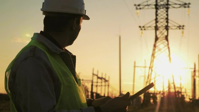Silhouette of engineer walk on field with electricity towers. Electrical engineer with high voltage electricity pylon at sunset background. Power workers at work concept.