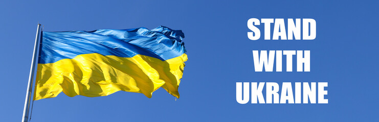 Large banner with the national flag of Ukraine flutters in the wind and text STAND WITH UKRAINE. The concept of protecting Ukraine from Russian aggression. War in Ukraine