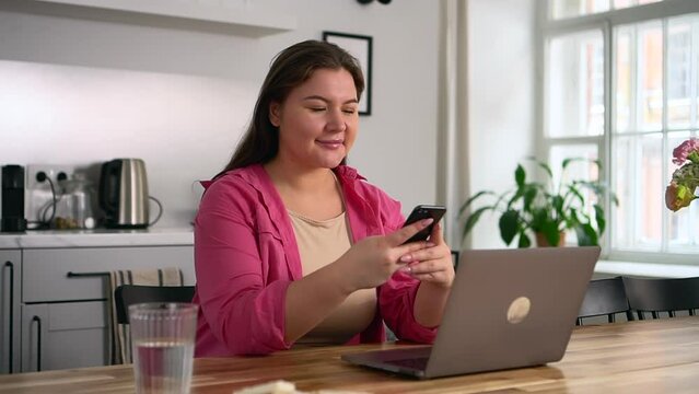 Young overweight businesswoman is holding phone and sitting at table with laptop in home spbd. Closeup view of beautiful oversize European woman holds smartphone in her hand and looks at display with