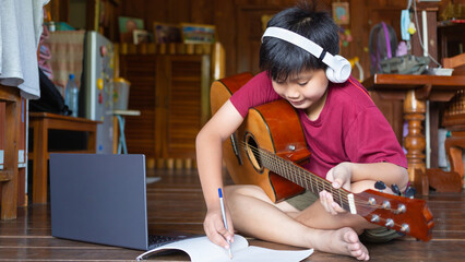 A cute Asian boy wearing headphones video calls learning  .music and writing songs online on his...