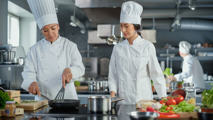 Restaurant Kitchen: Portrait of Asian and Black Female Chefs Preparing Dish, Cutting Vegetables, Stirring Ingredients on the Frying Pan. Two Professionals Cook Delicious, Authentic Food, Healthy Meals
