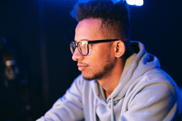 Dark skinned young man with a beaming smile, wears a gray parka, and glasses on dark background splashed by blue light