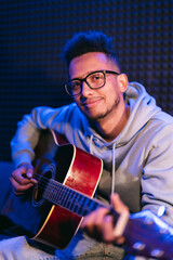 Young afro musician wearing glasses playing acoustic guitar, on dark background