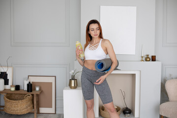 Young Caucasian woman in sportswear posing for camera with water bottle and yoga mat