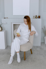 Woman in a white tracksuit sits on an armchair and watches the Internet on her phone.