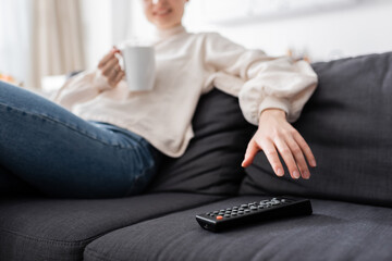 partial view of blurred woman with cup of tea near tv remote controller on couch