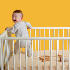 Happy infant baby boy is standing in a crib, yellow studio background. Smiling caucasian toddler kid in pajamas