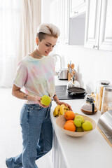 trendy young woman holding apple and bananas near bowl with fresh fruits in kitchen